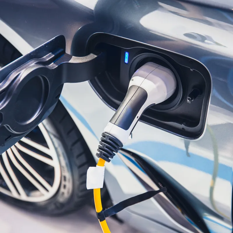 Close-up of an electric vehicle charging port with a charging connector plugged in, showing the cable, port door, and rear wheel.