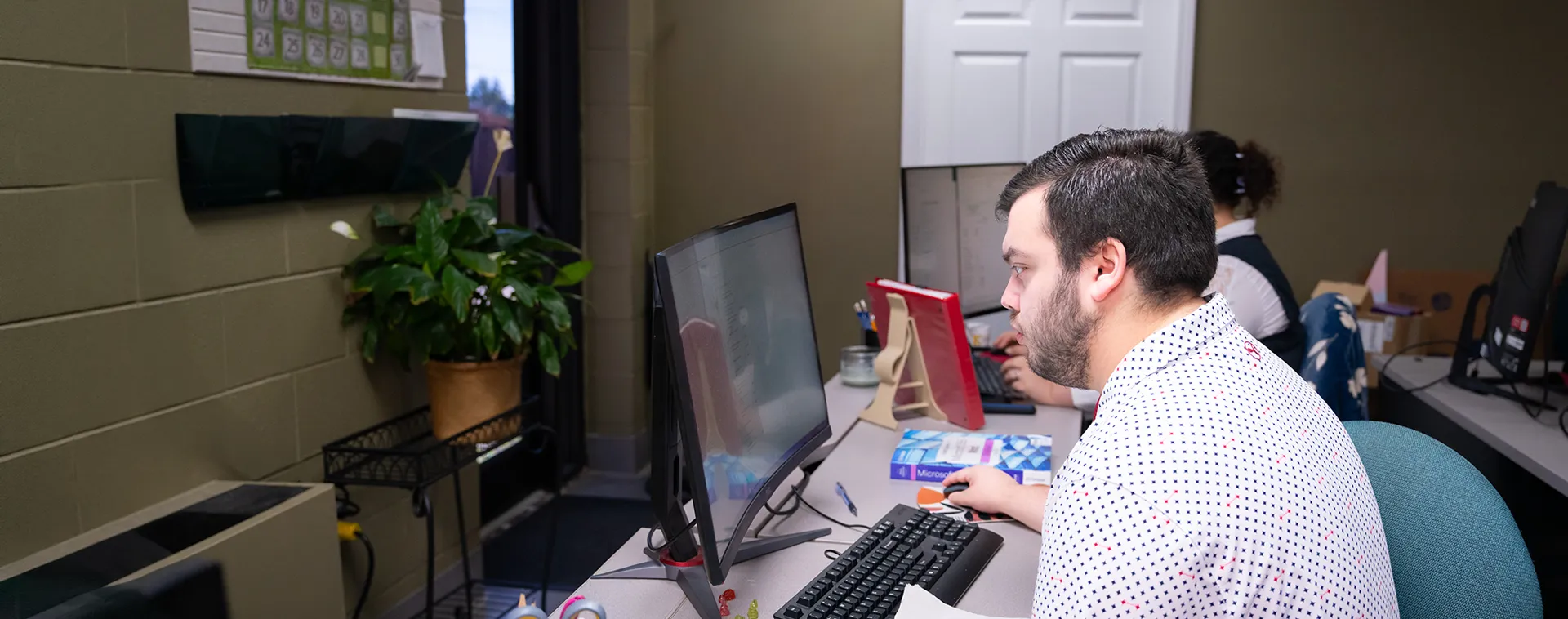 Student seated at a computer workstation in an office classroom, with a monitor, keyboard, books, and plants nearby.