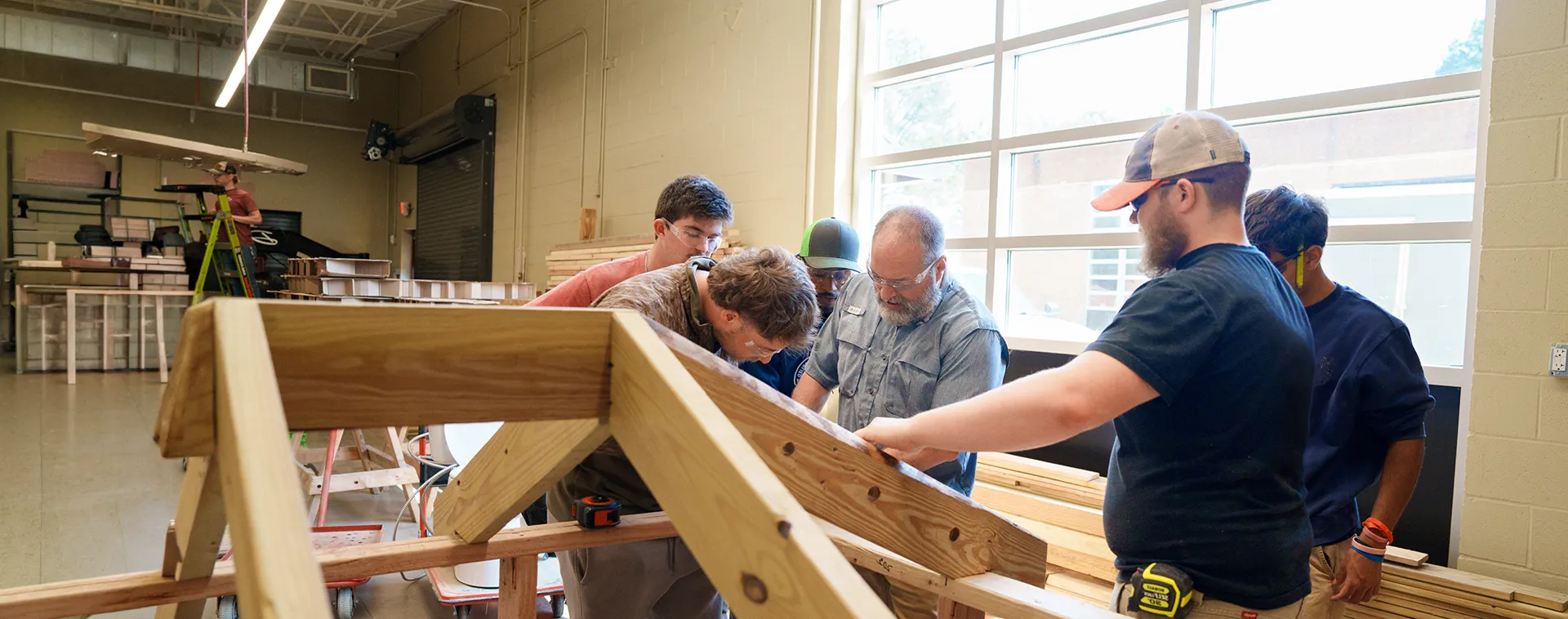 Group of students working together on a wooden roof frame inside a construction lab with lumber, tools, and large windows.