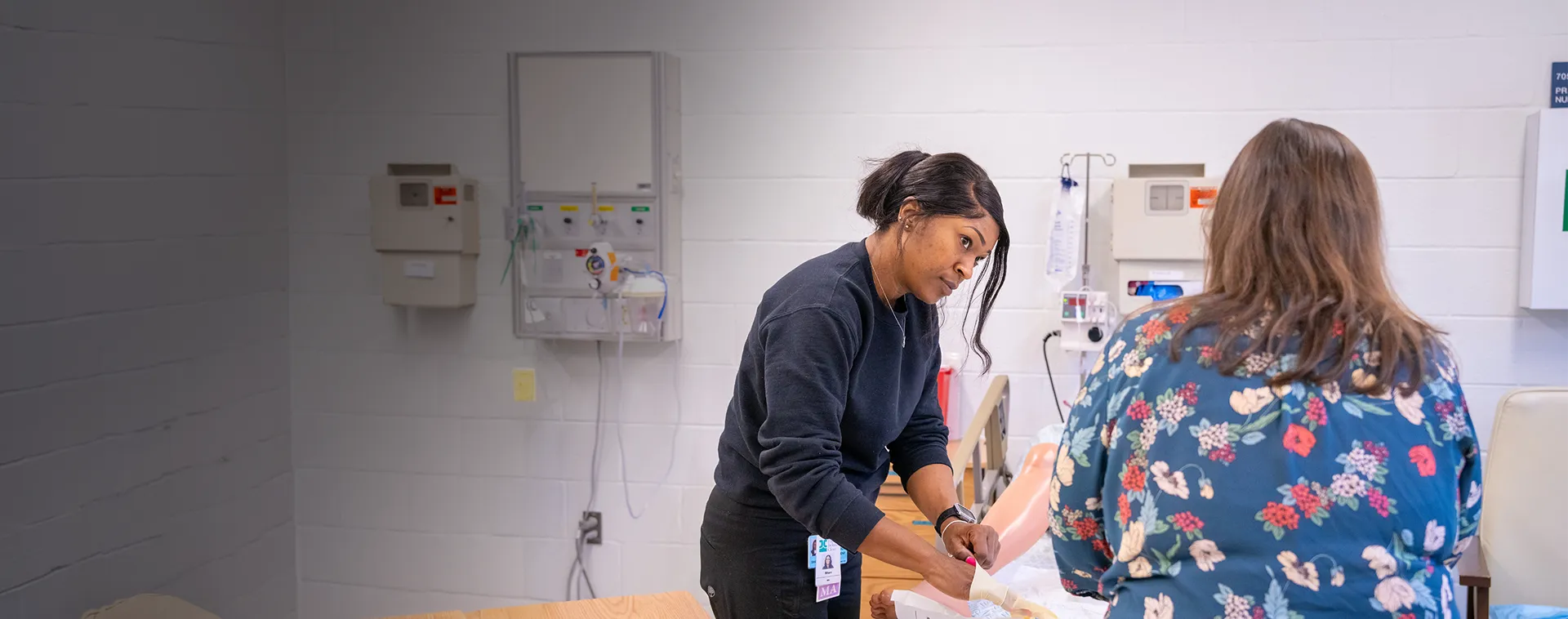 Person stands beside a hospital bed, assisting with care in a clinical skills lab with wall‑mounted medical equipment.
