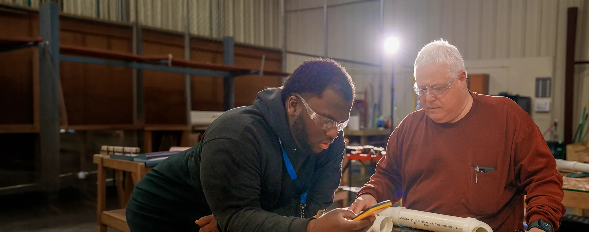 Two people review measurements on a PVC pipe at a workbench inside an industrial training lab with shelving and tools.