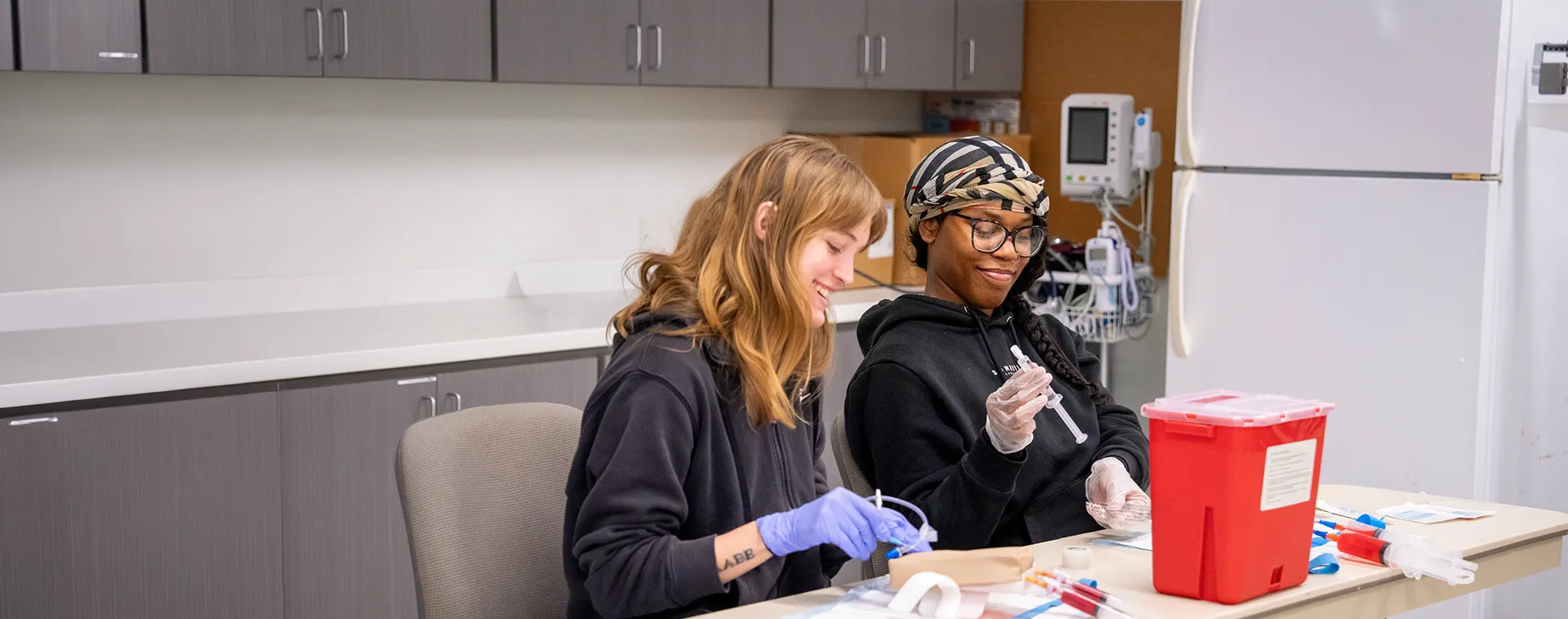 Two people wearing gloves sit at a table preparing syringes and medical supplies in a clinical training room.