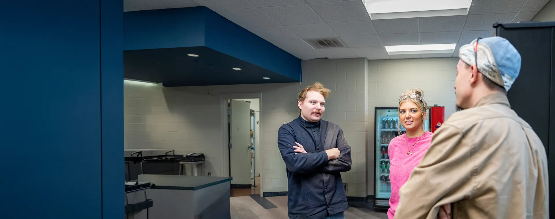 Three people standing and talking inside a campus common area with vending machines, counters, and overhead lighting.