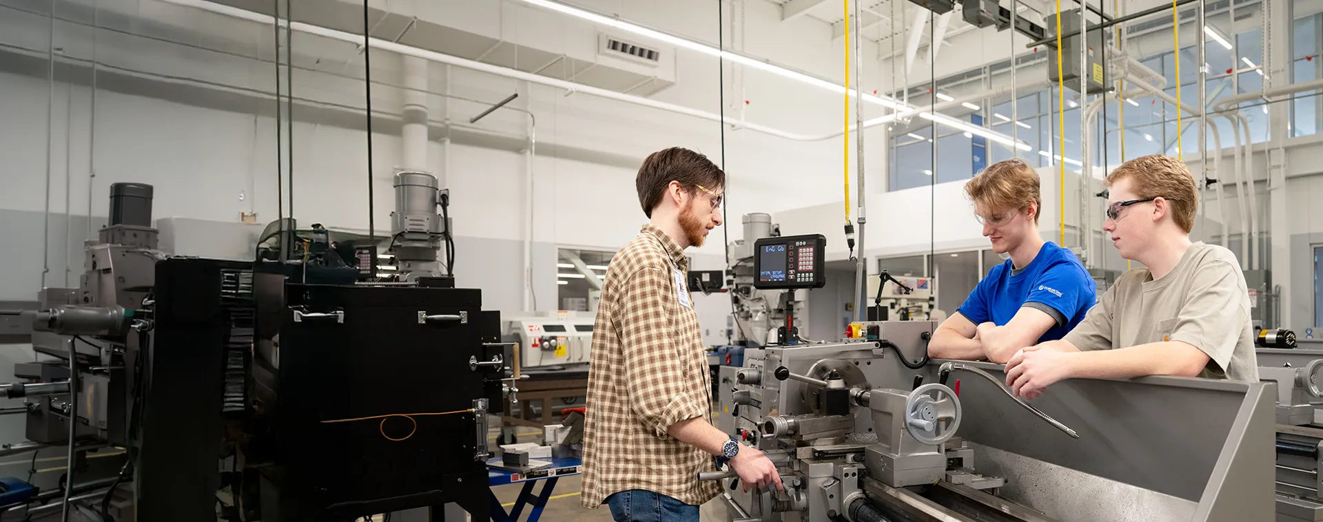 Three people stand around industrial machining equipment in a lab, with metal lathes, control panels, and overhead lighting.