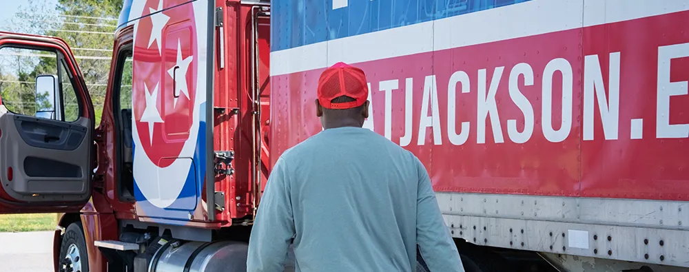 Person wearing a red cap stands beside a TCAT Jackson semi‑truck with an open cab door and large Jackson.edu logo.