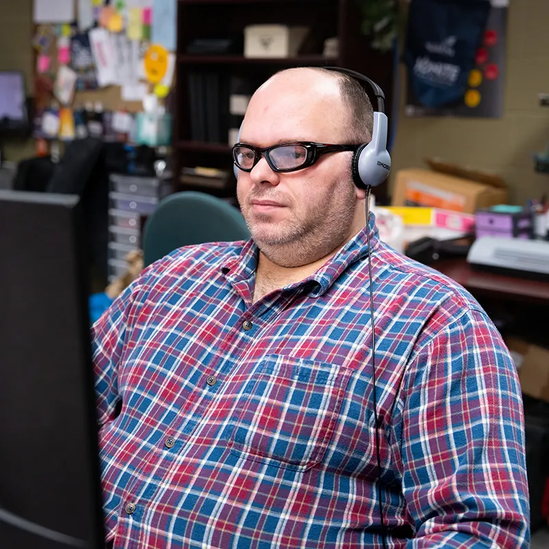 Student wearing headphones seated at a computer workstation in an office with shelves, boxes, and bulletin boards behind.