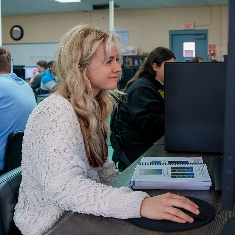 Student using a computer mouse at a desk with a printed workbook, with other students and monitors in the background.