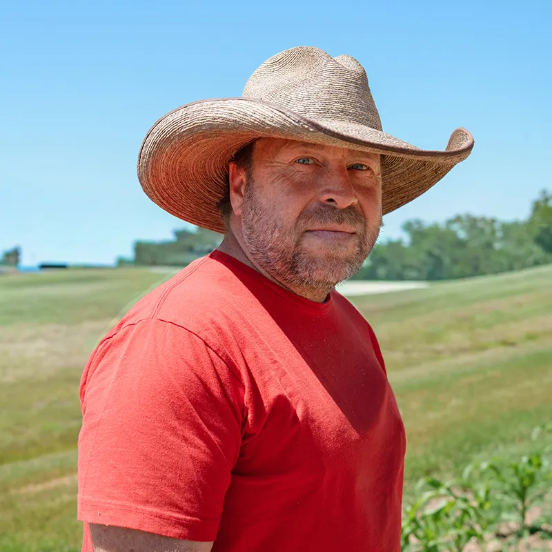 Person wearing a wide‑brim hat standing in a grassy field with rolling hills and trees in the background.