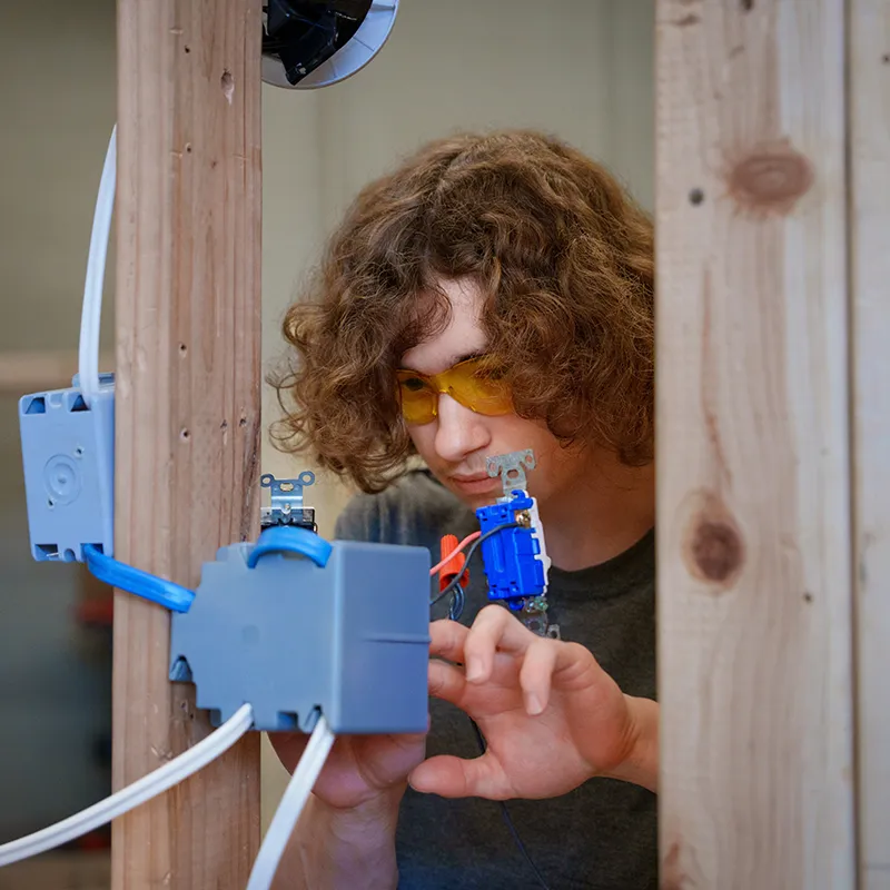 Student adjusting wires inside a blue electrical box mounted between wooden studs in an electrical training area.