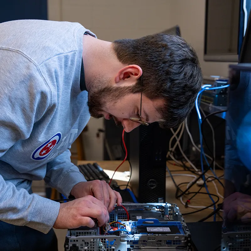 Student working inside an open desktop computer, connecting internal cables at a workstation with monitors nearby.