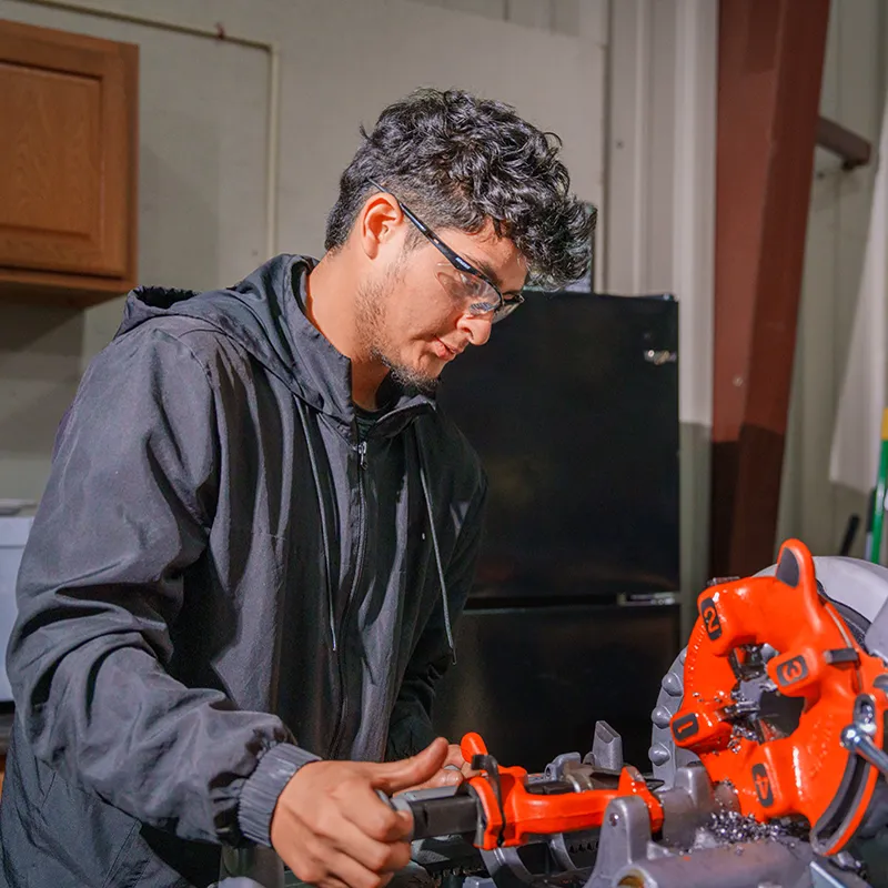 Person operates a pipe‑threading machine, holding a metal pipe in a workshop with tools and equipment nearby.