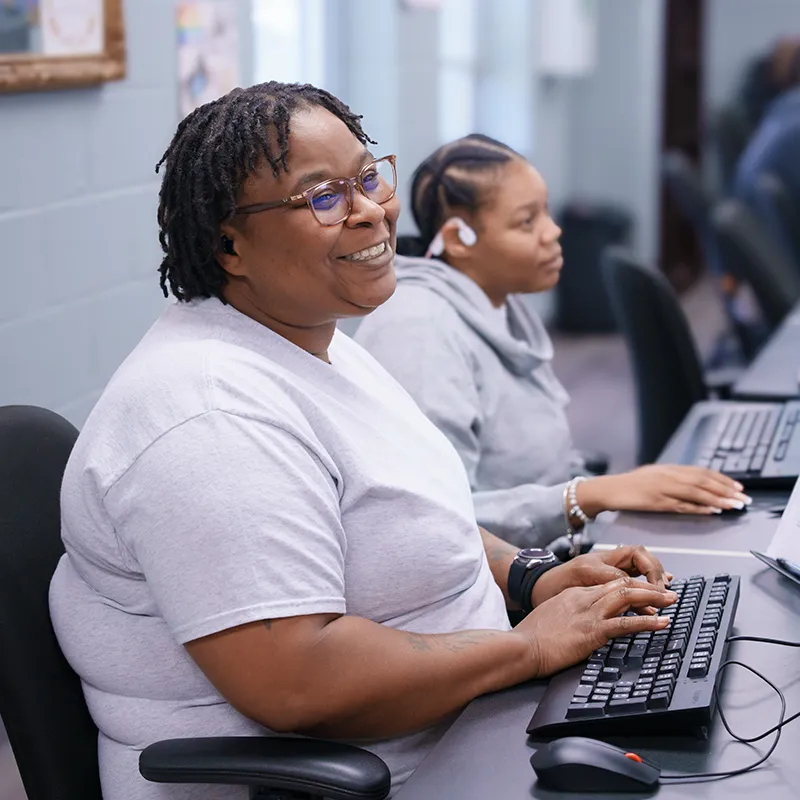 Two people sit at computers typing on keyboards in a classroom with rows of workstations