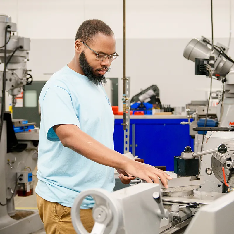 Person stands at a metal lathe, positioning a tool near a rotating metal piece in a workshop setting