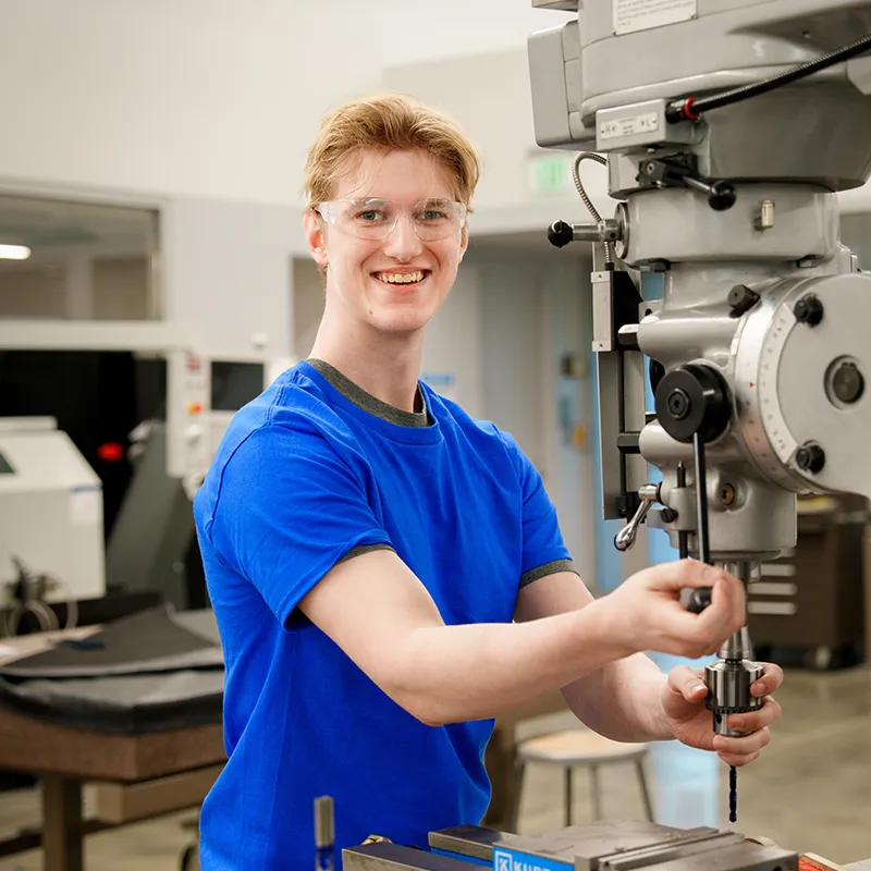 Person uses a drill press to lower a cutting tool onto a metal part in a machining training lab.