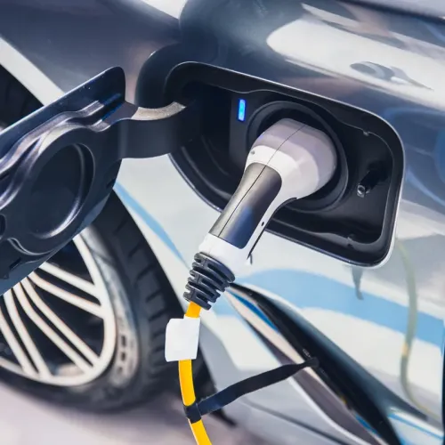Close-up of an electric vehicle charging port with a charging connector plugged in, showing the cable, port door, and rear wheel.