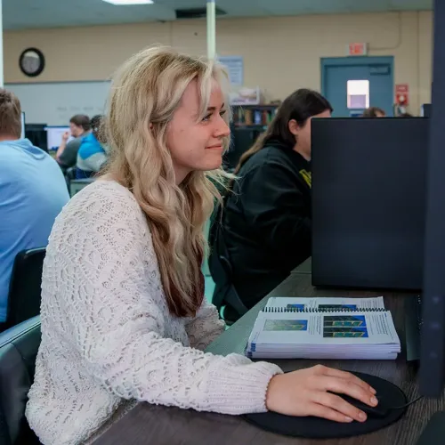 Student using a computer mouse at a desk with a printed workbook, with other students and monitors in the background.