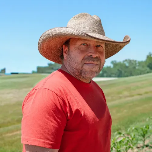Person wearing a wide‑brim hat standing in a grassy field with rolling hills and trees in the background.