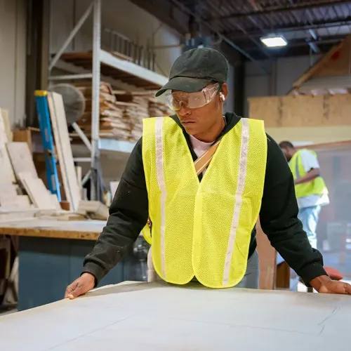 Student wearing a safety vest standing at a worktable with a large sheet of material in a woodworking shop.