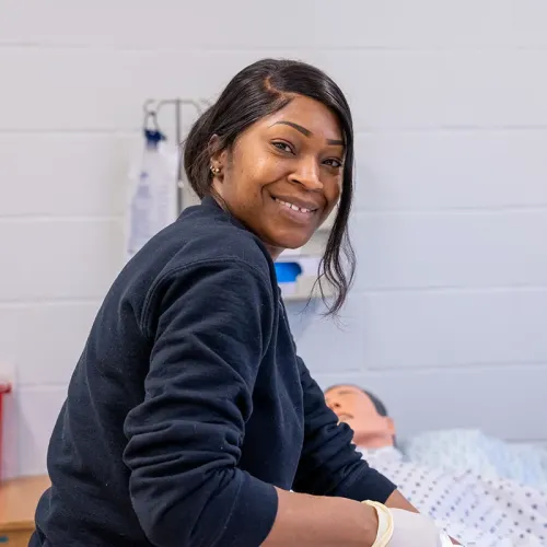 Person wearing gloves provides care to a medical mannequin in a hospital bed inside a training lab.