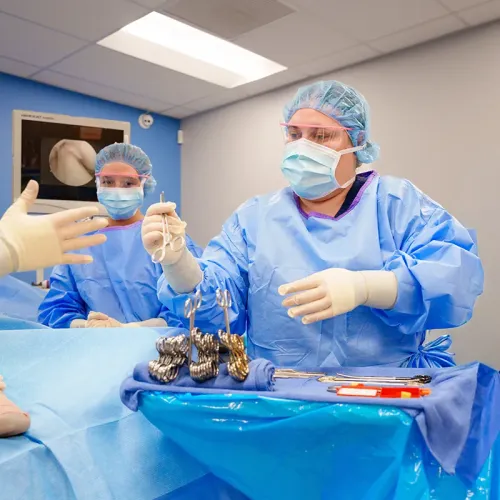 Two people in surgical gowns and gloves handle metal surgical instruments on a draped table in a training lab.