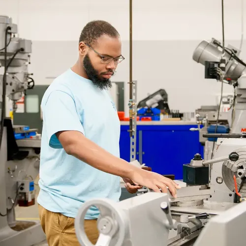 Person stands at a metal lathe, positioning a tool near a rotating metal piece in a workshop setting