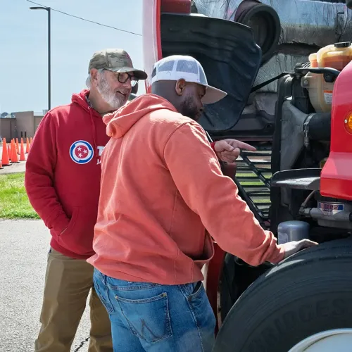 Two people stand beside a semi‑truck with the hood open, looking at engine components during hands‑on instruction.