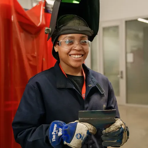Person wearing a welding helmet and gloves holds a metal plate inside a workshop with red safety curtains and equipment.