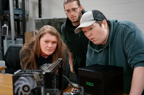 Three students gathered around a computer workstation, examining an open desktop computer case on a classroom table.