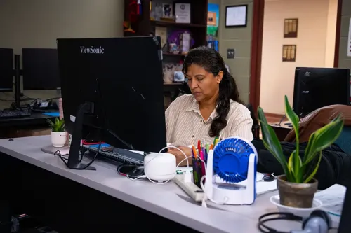 Student working at a front desk computer with office supplies, small plants, and additional monitors behind the counter.