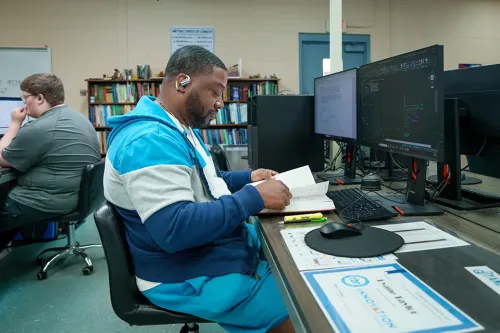 Student seated at a computer workstation reviewing a notebook beside multiple monitors in a classroom with bookshelves.