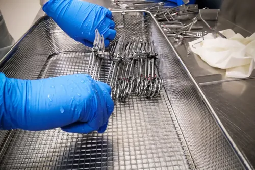 Gloved hands arranging metal surgical instruments in a wire basket on a stainless steel surface.