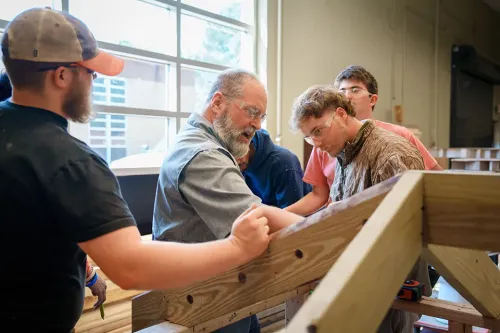 Students gathered around a partially built wooden structure, measuring and aligning boards in an indoor workshop.