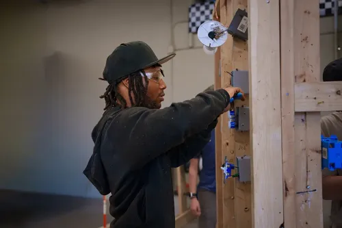 Student using a screwdriver to secure wiring inside an electrical box on a wooden training frame in a lab.
