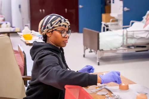 Person wearing gloves prepares medical supplies at a table, with a hospital bed and training room in the background.