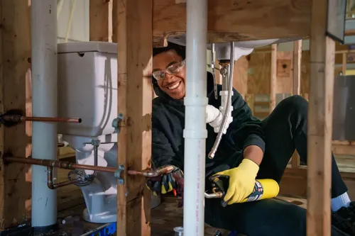 Person works beneath a framed structure, applying sealant to plumbing pipes near a toilet during hands‑on training.
