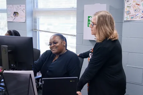 Person stands beside another seated at a computer, pointing and providing guidance in a classroom computer lab