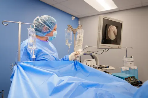 Person in surgical gown and gloves stands beside IV bags and medical equipment behind a blue surgical drape.