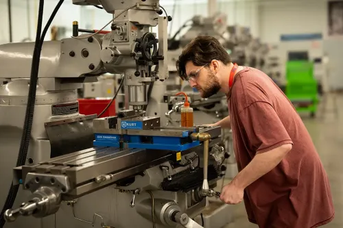 Person leans over a milling machine, adjusting a metal workpiece secured in a vise inside a machine shop.