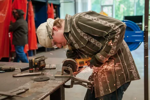 Person grinds a metal piece on a workbench, producing sparks, with welding tools and red safety curtains in the background.